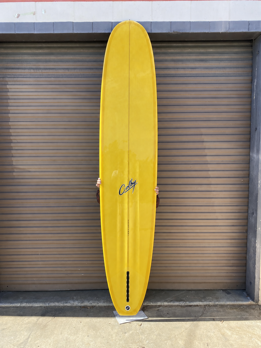 Golden yellow Colby Tick Bird longboard underside held upright in front of a grey roller shutter