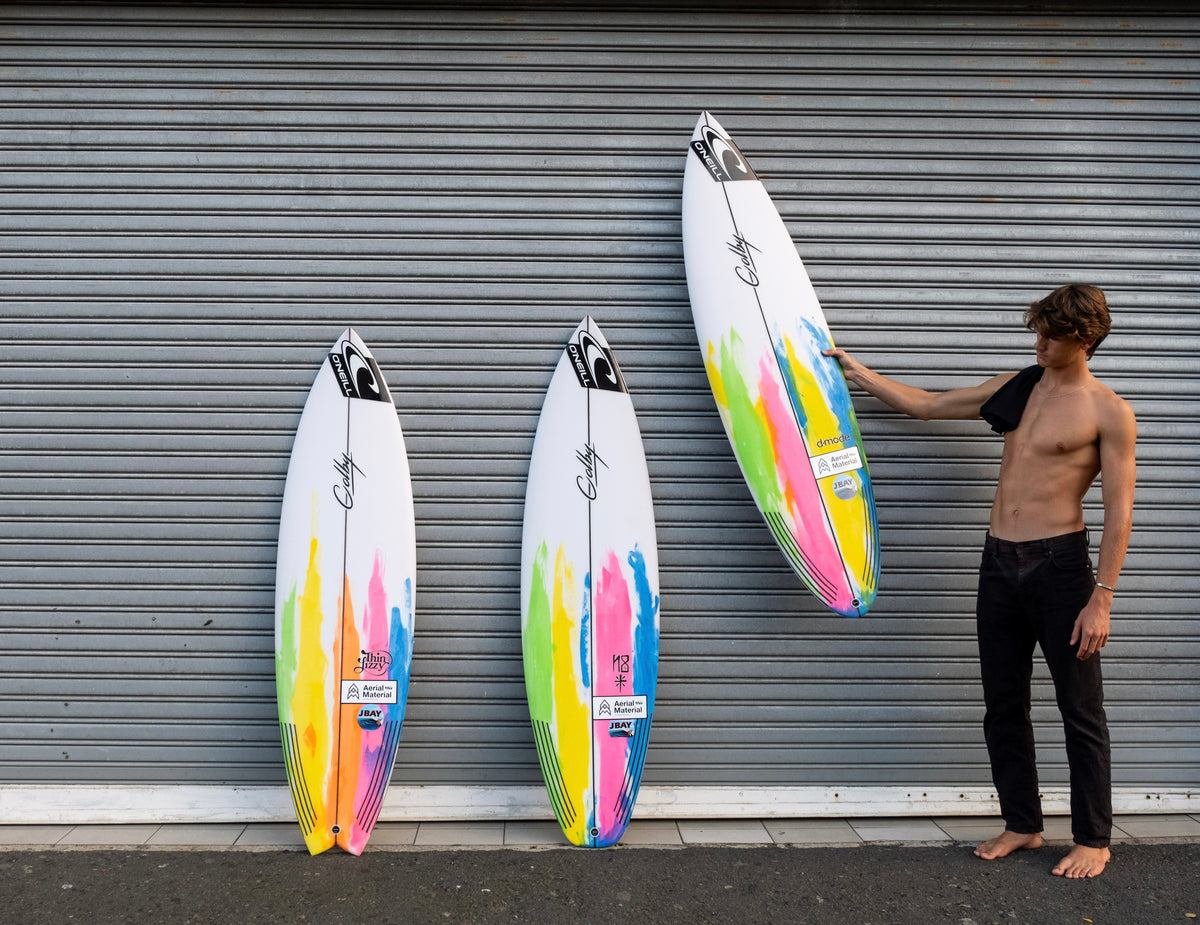 Three white surfboards with vibrant multicolor paint splatter designs are lined up against a gray metal garage door, with a shirtless man in black pants standing on the right and touching the tallest surfboard.